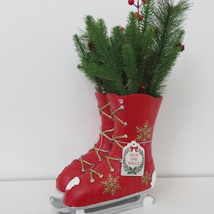May include: A red decorative ice skate planter filled with faux evergreen branches and red berries. The skates have gold-colored laces and snowflake accents. A tag reads "Deck the Halls."