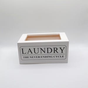 May include: White rectangular laundry box with a rectangular opening at the top. The box has the word "LAUNDRY" in a black, bold font, with "THE NEVER ENDING CYCLE" below it.