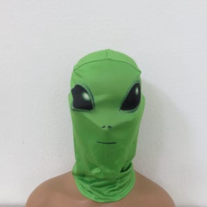 May include: A green alien mask with large black eyes and a small mouth. The mask is made of a soft, stretchy material and covers the entire head and neck. The mask is displayed on a mannequin.