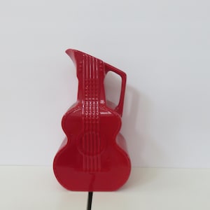 Könnte beinhalten: Ein glänzender roter Krug in Form einer Akustikgitarre. Der Krug hat einen Griff und einen Ausguss, und das Gitarrendesign umfasst die Saiten und das Schallloch. Der Krug ist ein dekorativer Gegenstand.