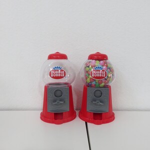 May include: Two red and clear plastic gumball machines with a silver base. Each machine has a Dubble Bubble logo and is filled with colorful gumballs.