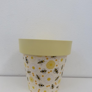 May include: A decorative plant pot with a bee and daisy design. The pot has a light yellow rim and a cylindrical shape. The pattern features black and yellow bees and white daisies on a cream background.