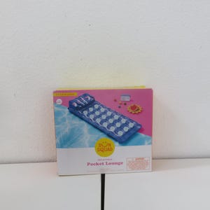 Puede incluir: Un Pocket Lounge inflable en caja de Sun Squad. El salón azul y blanco mide 1,83 metros de largo. La caja tiene un fondo rosa con un gráfico de piscina y un logotipo amarillo de Sun Squad.