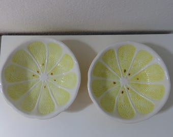 May include: Two white ceramic bowls, each shaped like a lemon slice. The bowls have a yellow interior with a textured design mimicking the fruit's segments and brown seed details. The bowls are on a white surface.