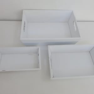 May include: Three white rectangular trays of varying sizes are arranged on a white surface. Each tray has a rectangular opening for a handle on each side. The trays are empty and appear to be made of wood or a similar material.