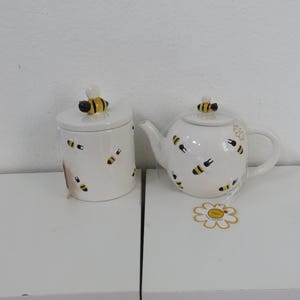 Puede incluir: Juego de tetera y azucarero de cerámica blanca decorado con motivos de abejas negras y amarillas. La tetera tiene asa y boquilla, y el azucarero tiene tapa. Ambos tienen asas con forma de abeja. Un juego de cocina decorativo con temática de abejas.