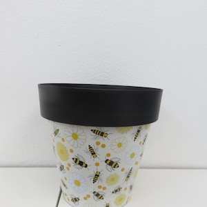 May include: A cylindrical flower pot with a black rim. The pot's body is white with a pattern of yellow daisies and black and yellow bees. The pot is sitting on a white surface.