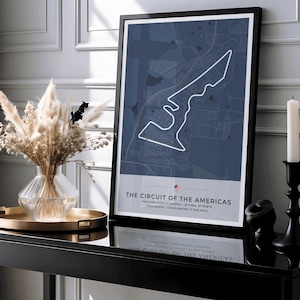 May include: A framed poster showing the Circuit of the Americas race track in white against a navy blue backdrop. The text "THE CIRCUIT OF THE AMERICAS" and track details are at the bottom. The poster is displayed on a black table, with a vase and candle.