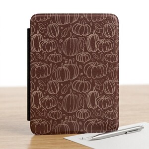 May include: A rectangular e-reader case featuring a dark brown background with a repeating pattern of light brown line-art pumpkins, leaves, and small acorns. The case has a textured surface and a black spine visible on the left side of the device.