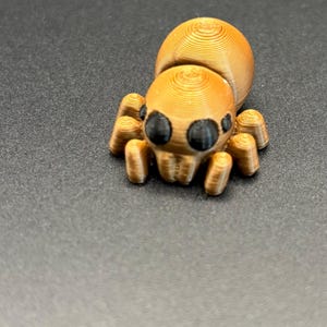 May include: A 3D-printed spider figurine in shades of gold and brown, with black eyes. The spider has a segmented body and legs, and is set against a dark background. The figurine is a decorative item.