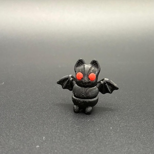 May include: A small, black bat figurine with red eyes and outstretched wings. The bat has a detailed, textured body and small, pointed ears. The wings are also textured and spread out to the sides. The figurine is standing upright.