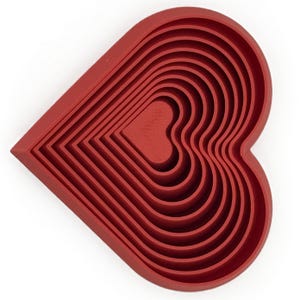 May include: A nested set of red heart-shaped cookie cutters. The cutters are arranged in a layered design, with each heart slightly smaller than the last. The color is a deep, solid red, and the cutters appear to be made of a flexible material.