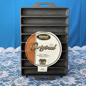 May include: A cast iron Wagner 1891 Original loaf pan with a rectangular shape and multiple individual loaf molds. The pan has a handle on each end and a circular label with the Wagner logo and "Original" text. The pan is displayed on a black metal stand.