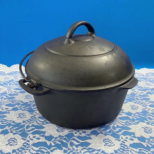 May include: A black cast iron Dutch oven with a lid and handle. The pot is round with a handle on the lid and two handles on the sides. The pot is sitting on a white lace tablecloth with a blue background.