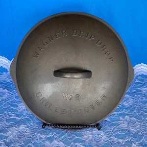 May include: A vintage Wagner Drip Drop skillet cover, size No. 9, made of cast iron. The lid has a handle and is displayed on a black metal stand. The text "WAGNER DRIP DROP" and "SKILLET COVER" are embossed on the lid.