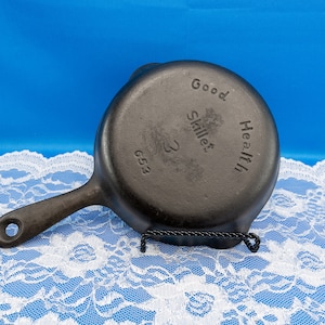May include: A small, black cast iron skillet with a single handle and a hole for hanging. The words "Good Health Skillet 3 653" are embossed on the bottom. The skillet is displayed on a white lace tablecloth against a blue background.