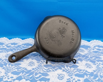 Puede incluir: Una pequeña sartén de hierro fundido negro con un solo mango y un agujero para colgar. Las palabras "Good Health Skillet 3 653" están grabadas en la parte inferior. La sartén se muestra sobre un mantel de encaje blanco sobre un fondo azul.