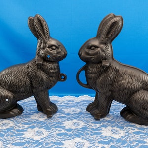May include: Two black rabbit-shaped molds for making chocolate or other treats. The molds are made of metal and have a textured surface. They are sitting on a white lace tablecloth.