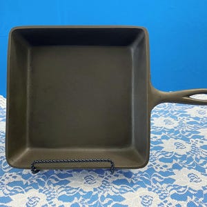 May include: A square, dark gray cast iron skillet with a handle. The pan is sitting on a black metal stand, against a blue background. The handle has a hole for hanging. The pan is approximately 10 inches square.