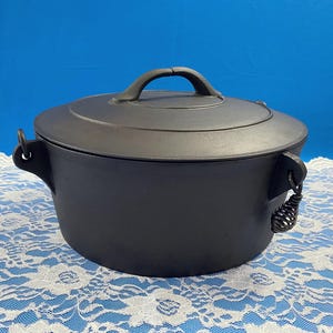 May include: A black cast iron Dutch oven with a lid and handle. The pot has two side handles and a handle on the lid. The Dutch oven is sitting on a white lace tablecloth against a blue background.