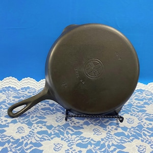 May include: A dark gray cast iron skillet with a long handle and a circular cooking surface. The bottom of the skillet features a logo and text. The skillet is displayed on a black metal stand against a blue background.