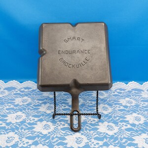 May include: A cast iron griddle with the text "SMART ENDURANCE BROCKVILLE" is displayed on a black metal stand. The griddle is square with a handle and a textured surface. The background is blue.