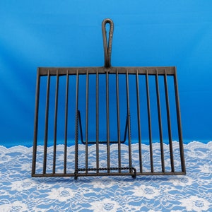 May include: Black metal grilling rack with a handle. The rack has 12 vertical bars and a stand with a decorative design.