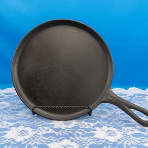 May include: A black cast iron skillet with a round cooking surface and a long handle. The skillet is sitting on a white lace tablecloth.