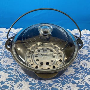 May include: A black cast iron pot with a clear glass lid and a metal handle. The pot has a stainless steel steamer insert. The lid has a clear glass knob. The pot is sitting on a white lace tablecloth.