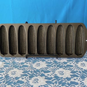 May include: A cast iron corn stick pan with eight corn cob molds. The pan is dark gray and has a handle on each end. The molds are detailed to resemble corn cobs. Perfect for baking corn bread.