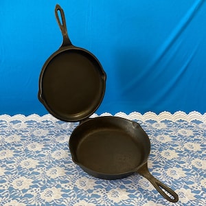 May include: Two black cast iron skillets with handles. The skillets are round with a dark, textured surface. One skillet is positioned above the other, with a blue background and a white lace tablecloth.