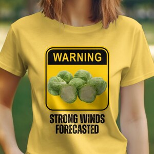 May include: Yellow t-shirt with a warning sign graphic. The sign is yellow with black text and a picture of brussels sprouts. The text on the sign reads "WARNING STRONG WINDS FORECASTED".