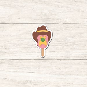 May include: A pink popsicle with a brown cowboy hat and a yellow and green face. The popsicle is on a stick.