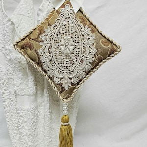 May include: A decorative, diamond-shaped ornament with a gold and brown patterned fabric, white lace overlay, and a white bow. It features a gold tassel and a braided gold cord for hanging. The ornament is approximately 10 cm wide.
