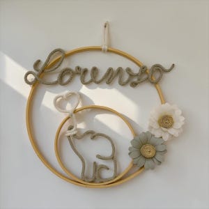 Pode incluir: Decoração de parede artesanal para berçário com o nome "Lorenzo" em fonte cursiva. O design inclui um aro de madeira, um elefante e duas flores de tecido em tons neutros. Projetado para ser pendurado na parede.