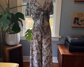 May include: A two-piece outfit on a mannequin. The set includes a short-sleeved top tied at the waist and wide-leg pants. Both pieces are a taupe color with a white floral pattern. The outfit is displayed in front of a window.