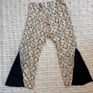 May include: A pair of wide-leg pants with a floral pattern in yellow, pink, and black on a white background. The pants have black panels on the lower legs. The pants are made of a soft, lightweight fabric.