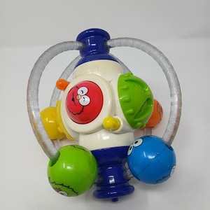 May include: A colorful baby rattle toy with a central white body, blue and yellow accents, and a clear ring. The rattle features a red circle with a cartoon character face, green and blue balls.