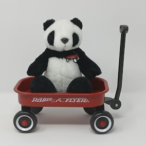 May include: A plush panda bear with black and white fur sits in a red Radio Flyer wagon. The wagon has black wheels with red centers and a black handle. The wagon has the words "Radio Flyer" printed on the side.