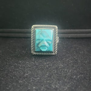 May include: A square turquoise ring with a silver setting. The ring has a rope-like design around the edge of the setting.
