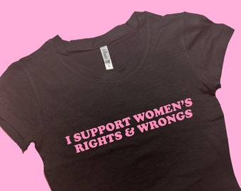 Puede incluir: Un top corto negro con el texto rosa "I SUPPORT WOMEN'S RIGHTS & WRONGS". La camiseta tiene cuello redondo y mangas cortas. El fondo es de color rosa liso.