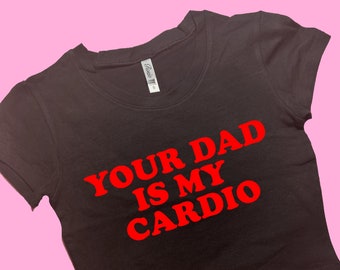 May include: Black crop top with the red text "YOUR DAD IS MY CARDIO". The shirt has a scoop neck and short sleeves. The background is a solid pink colour.