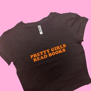 May include: A black crop top with the text "PRETTY GIRLS READ BOOKS" in orange lettering. The shirt has short sleeves and a scoop neck. The shirt is displayed on a pink background.