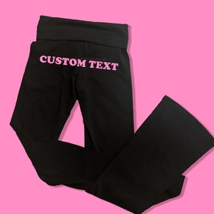 May include: Black yoga pants with a wide waistband and a flared leg. The pants have the text "CUSTOM TEXT" printed on the back in pink.