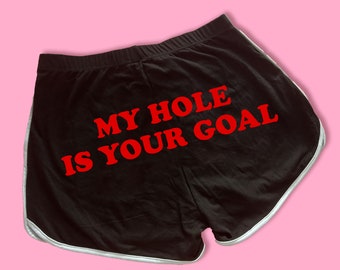 May include: Black athletic shorts with a white trim. The shorts have the text "MY HOLE IS YOUR GOAL" printed in red.