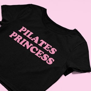 Puede incluir: Camiseta negra de corte corto con texto rosa que dice "PILATES PRINCESS"
