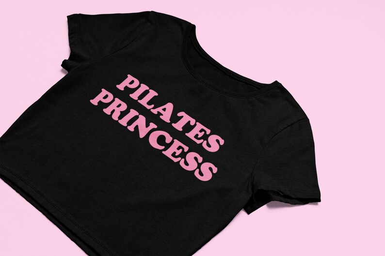 Puede incluir: Camiseta negra de corte corto con texto rosa que dice "PILATES PRINCESS"