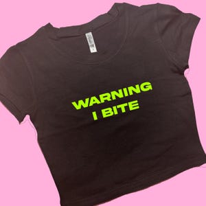 Könnte beinhalten: Ein schwarzes Crop-Top mit dem neongrünen Text "WARNING I BITE". Das Shirt hat kurze Ärmel und einen Rundhalsausschnitt. Der Hintergrund ist einfarbig rosa.