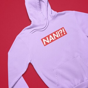 K&ouml;nnte beinhalten: Ein helllila Kapuzenpullover mit einer Vordertasche und einer Kapuze mit Kordelzug. Der Kapuzenpullover zeigt das Wort "NANI?!" in wei&szlig;er Schrift in einem roten Rechteck auf der Brust.