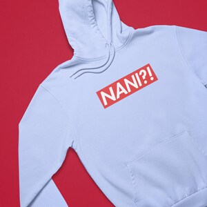 K&ouml;nnte beinhalten: Hellblauer Kapuzenpullover mit Fronttasche und Kordelzugkapuze. Auf der Vorderseite des Hoodies steht das Wort "NANI?!" in wei&szlig;er Schrift in einem roten Rechteck.